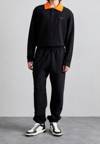 Polo a maniche lunghe nera con colletto arancione, abbinata a pantaloni della tuta neri e sneakers bianco e nere. Presenta una chiusura a bottoni e una vita con cordino.