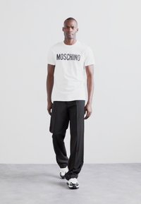 Tricou alb din bumbac cu un logo negru "MOSCHINO", asortat cu pantaloni negri, croiți și teniși alb-negru. Fundal simplu.