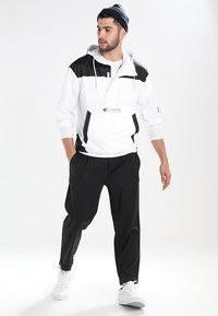 Hvid pulloverjakke med sorte detaljer, justerbar hætte og frontlomme; parret med løstsiddende sorte bukser og hvide sneakers.