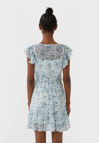 Femme à la peau foncée et aux cheveux en chignon, portant une robe courte blanche à motifs floraux avec des fleurs bleues et vertes, des manches volantées et une taille élastique, vue de dos.