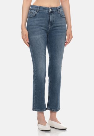 Donna che indossa jeans blu a vita media e gamba dritta e scarpe piatte bianche, in piedi davanti a uno sfondo chiaro e uniforme.