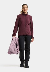 Woman standing wearing a maroon zip-up fleece jacket, black pants, black hiking shoes, holding a folded light purple jacket in one hand.