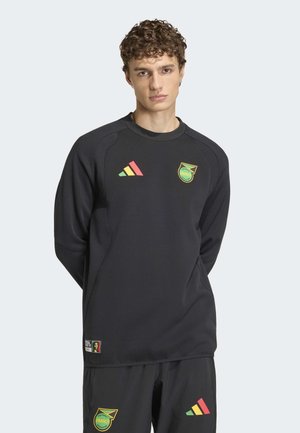 Joven con sudadera negra de fútbol de Jamaica y pantalones cortos a juego con logotipos de Adidas en rojo, amarillo y verde sobre fondo liso.