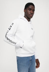 Hoodie branco em tecido suave, com um bolso frontal e capuz com cordão. Logótipos pretos impressos na manga e no peito. Corte casual.
