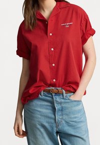 Red short-sleeve button-up shirt with a collar and embroidered "POLO SPORT" logo. Worn over light blue jeans, featuring a braided belt.