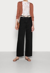 Jeans neri a gamba larga, cintura beige, t-shirt bianca e tie-dye, cardigan color ruggine, borsa a tracolla marrone e sandali scivolosi color beige.
