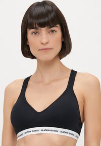 Melnā sporta krūšturiņa ar V-veida kakla izgriezumu, regulējamiem siksniņām un baltu zīmola elastīgo joslu apakšā. Gluda tekstūra un minimālisma stils.