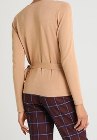 Haut beige à manches longues avec une taille ceinturée, un décolleté rond et une texture lisse. Associé à un pantalon à carreaux marron avec des accents bleus.