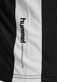 Sort atletisk stof med en hvid sideløsning, der har den prægede tekst "hummel PERFORMANCE" i sort langs sømmen.
