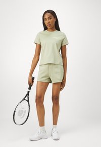 T-shirt verde claro de mangas curtas e calções combinando, feitos de tecido macio. Usado com ténis brancos, segurando uma raquete de ténis preta.