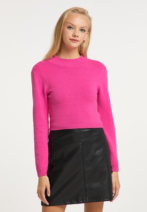 Młoda kobieta z długimi blond włosami, ubrana w jasnoróżowy sweter i czarną skórzaną spódnicę, stoi na jednolitym tle.