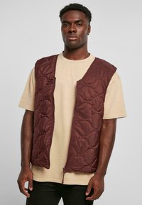 Bordeauxfärgad vadderad väst, ärmlös med en dragkedja fram och en mjuk yta, bärs över en beige t-shirt. Enkel, vadderad design utan mönster.