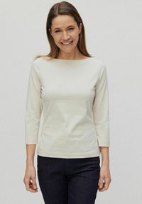 Gräddfärgad långärmad topp med båturringning, figursydd midja och mjuk textur, kombinerad med mörka jeans.