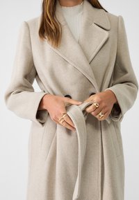 Beige wollen blend jas met een dubbelgeknopte ontwerp, inkeping-lapels en een strikriem. Gladde textuur met subtiele patroon details.