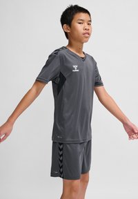 Graues Sportoutfit mit einem kurzärmligen V-Ausschnitt-Jersey und passenden Shorts. Die Textur ist glatt; die Shorts zeigen ein schwarzes Zickzackmuster.