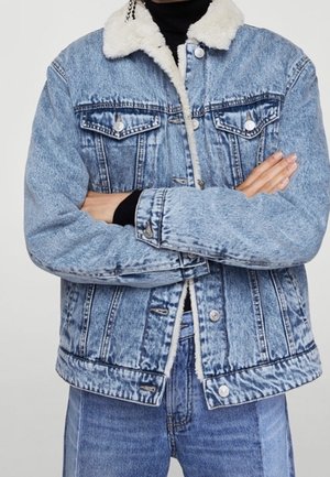 Persoon die lichtblauwe spijkerjas draagt met witte sherpa kraag en voering, gecombineerd met bijpassende spijkerbroek en zwarte coltrui.