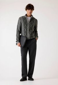 Maglione grigio lavorato a maglia con motivi a diamante, pantaloni neri e mocassini; tiene una custodia per laptop nera. Design semplice e strutturato.