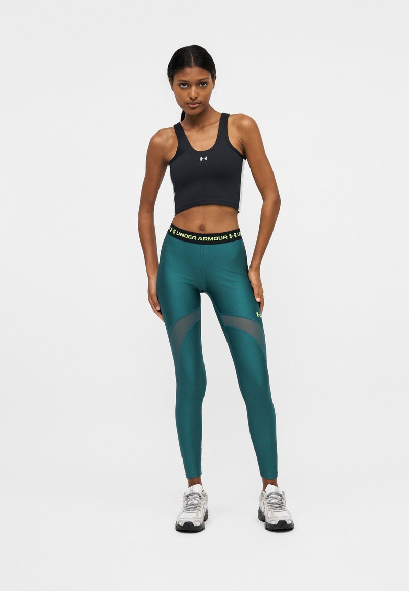 Mujer joven de pie, vestida con un top corto negro de Under Armour, mallas azul verdosas con cintura con logo y zapatillas blancas sobre fondo liso.