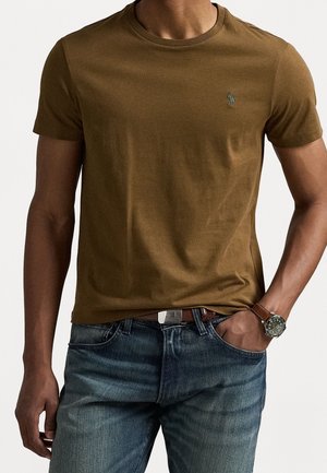 Homme portant un t-shirt à manches courtes vert olive avec un logo brodé, un jean bleu, une ceinture marron et une montre-bracelet, une main dans la poche.