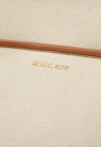 Tissu beige texturé avec un motif perforé léger et des accents en cuir marron. Logo "Michael Kors" doré près du bas.