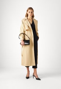 Béžový trench coat s knoflíkovými detaily, vrstveným designem a dlouhými rukávy, doplněný černou kabelkou a černými špičatými podpatky.
