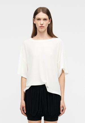 Femme aux cheveux bruns lisses portant un haut blanc ample à manches courtes et un short noir froncé, debout devant un fond clair uni.