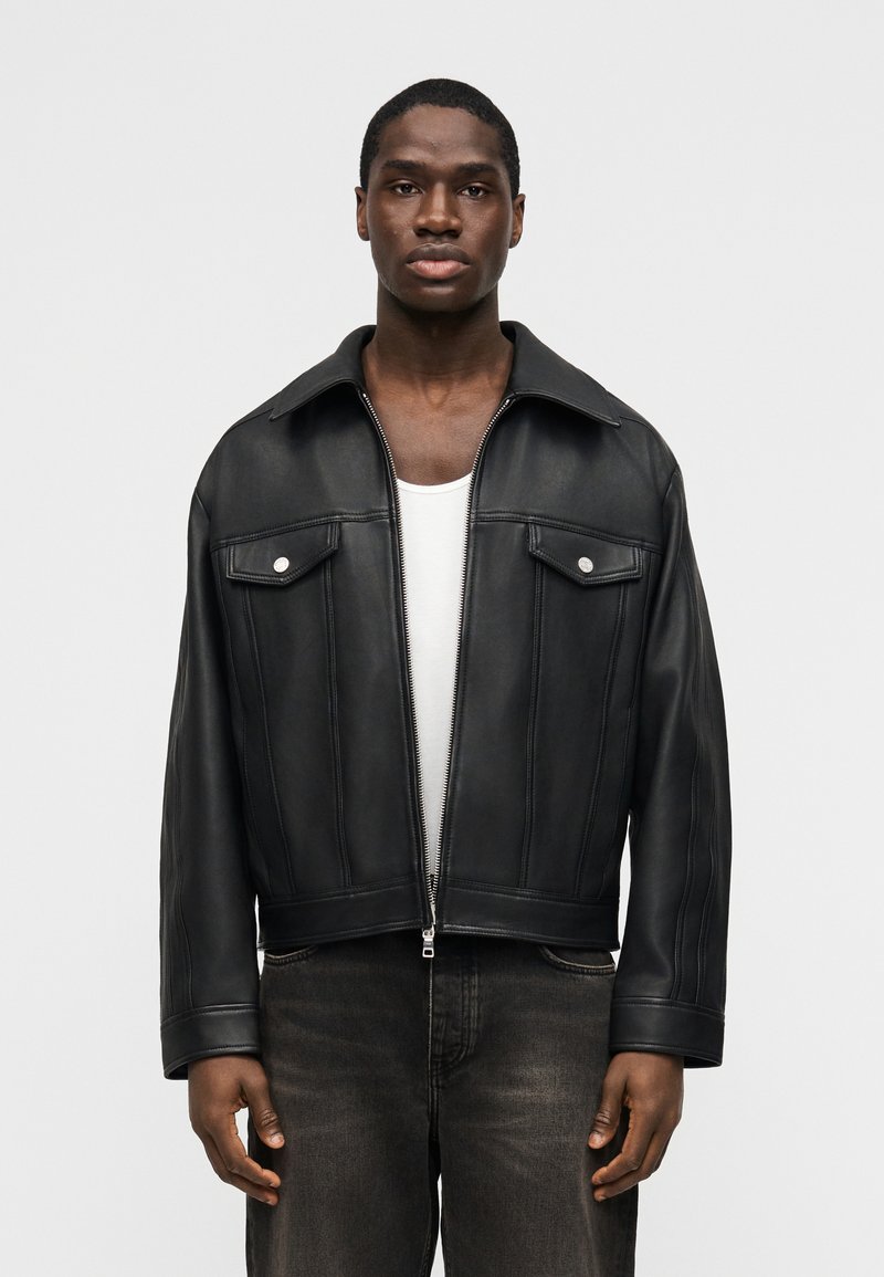 Άνδρας που φοράει μαύρο δερμάτινο  μπουφάν πάνω από λευκό τοπ χωρίς μανίκια και ξεθωριασμένο μαύρο τζιν, όρθιος μπροστά σε απλό φωτεινό φόντο.