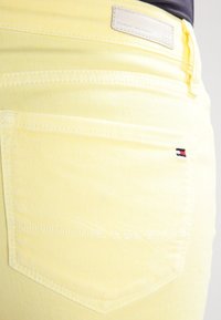 Jean en denim jaune avec une poche arrière ornée d'un design cousu et d'une petite étiquette logo aux couleurs rouge, blanc et bleu. Détails de couture visibles.