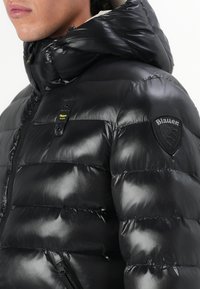 Schwarze Daunenjacke mit Kapuze, mit glänzender Textur, gestepptem Design und einem Patch-Logo am Oberarm. Reißverschluss-Tasche an der Seite.