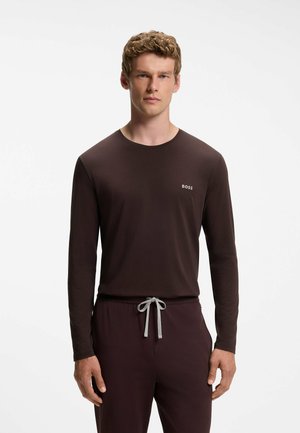 Jonge man met krullend haar die een donkerbruin shirt met lange mouwen en bijpassende broek met een grijs trekkoord draagt, staand tegen een witte achtergrond.