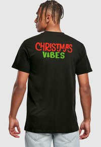 Black cotton T-shirt featuring the text "CHRISTMAS VIBES" in red and green with a Christmas tree icon, seen from the back.
