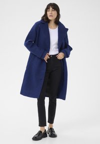 Blå ullblandad oversized kappa med en texturerad yta, breda slag och två framknappar, i kombination med svarta byxor och skinande loafers.