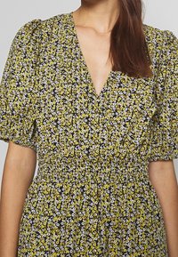 Květinové šaty s černým pozadím a žlutými a bílými květy, s výstřihem do V a balónovými rukávy. Nařasený pas zvýrazňuje postavu.