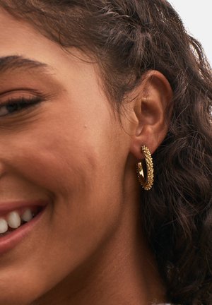 Orecchino ad anello in oro con superficie texturizzata e piccoli accenti decorativi, indossato all'orecchio di un modello. Forma curvata, finitura riflettente.