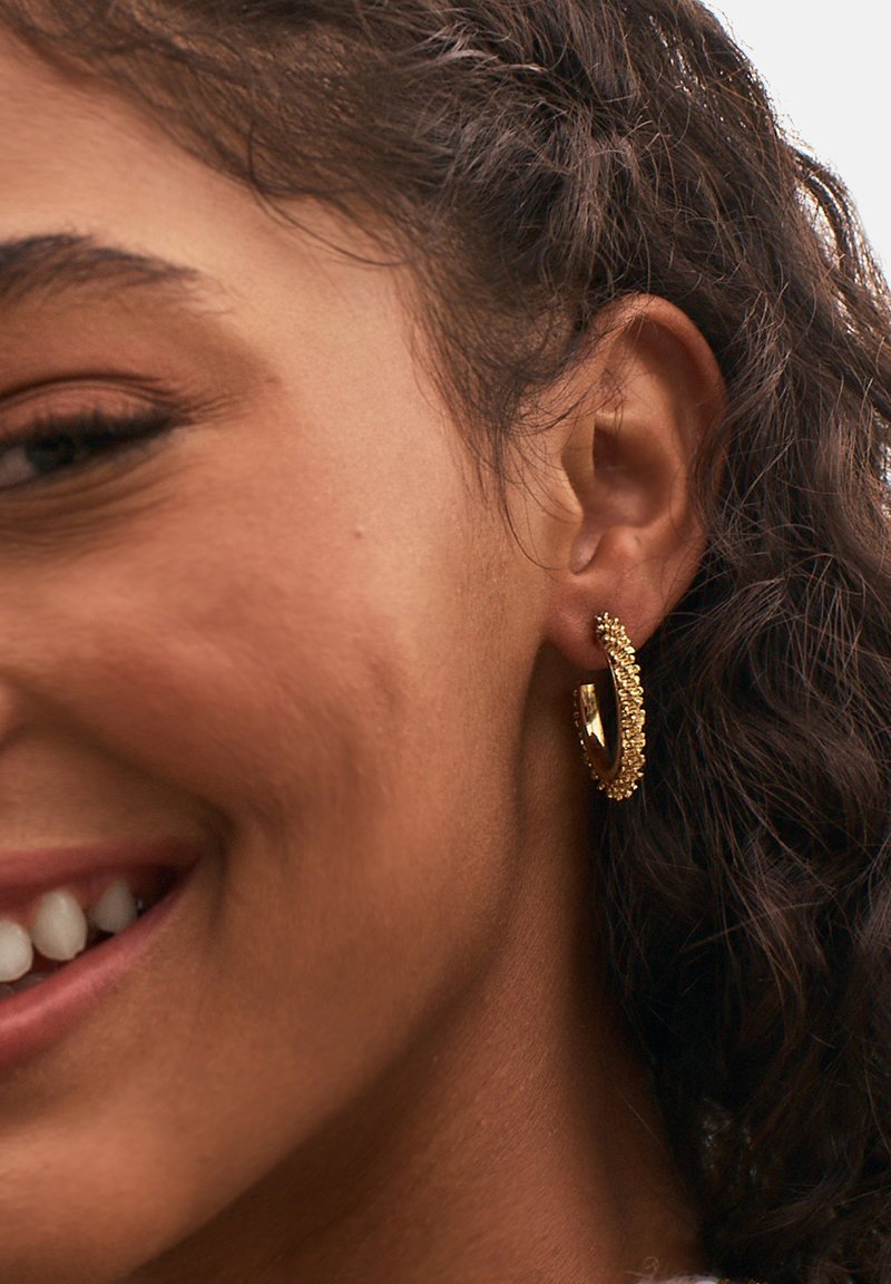 Orecchino ad anello in oro con superficie texturizzata e piccoli accenti decorativi, indossato all'orecchio di un modello. Forma curvata, finitura riflettente.