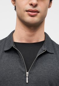 Γκρι πόλο με φερμουάρ και γιακά, με μεταλλικό φερμουάρ. Φοριέται πάνω από μαύρο πουκάμισο, με υφή υφάσματος και μοντέρνο σχεδιασμό.