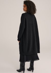 Lång svart cardigan med texturerat avslut, med sjunkna axlar och en lös passform, bärs över en plisserad svart kjol och knähöga svarta stövlar.