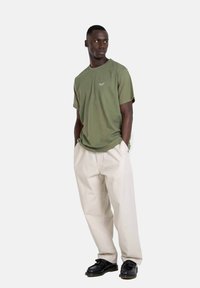 Olive green cotton t-shirt with short sleeves, featuring a small white logo. Paired with loose-fitting cream trousers and black shoes.