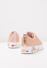 Las zapatillas de glitter en color oro rosa presentan un acento metálico en el talón, una suela blanca y un detalle de diseño floral cerca de la base.