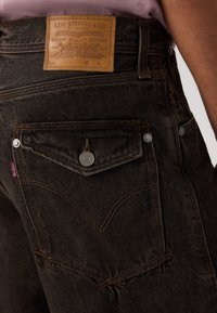 Κοντινή λήψη της πίσω τσέπης σκούρου τζιν Levi's με κουμπωτό καπάκι και δερμάτινη ετικέτα Levi Strauss & Co. πάνω από τη μέση.