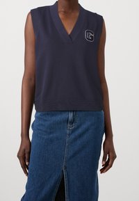 Námořnicky modrý tílko s výstřihem do V a vyšívaným písmenem "G" na hrudi. Vyrobeno z měkké, elastické látky.