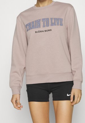 Persoon draagt een mauve sweatshirt met de tekst "TRAIN TO LIVE BJÖRN BORG" en zwarte Nike shorts met een wit swoosh-logo.
