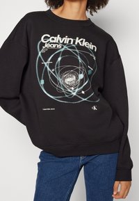 Černá mikina s grafickým motivem atomů a planet, s nápisem "Calvin Klein Jeans" v bílé a tyrkysové barvě. Měkká látka, volný střih.