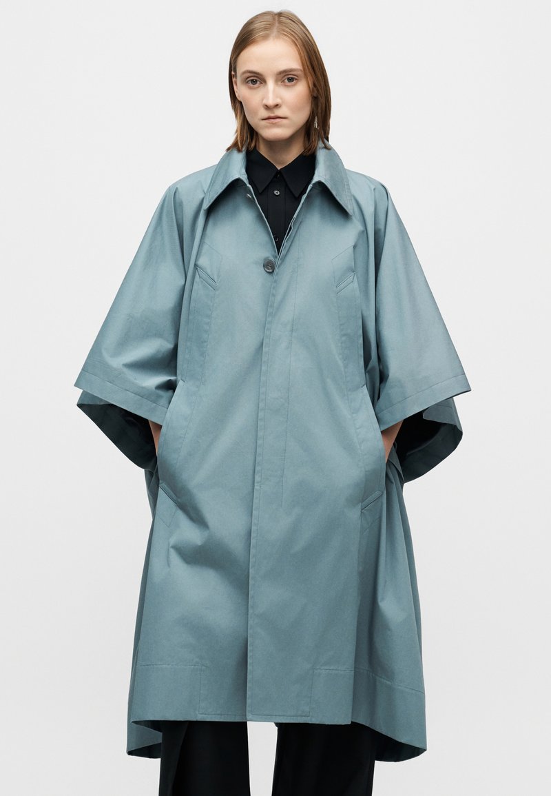 Manteau oversize bleu clair en tissu lisse, doté d'un col pointu, d'une fermeture boutonnée à l'avant et de grandes poches. Manches larges et courtes.