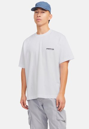 Camiseta blanca de algodón con un logo negro en el lado izquierdo del pecho, combinada con una gorra azul clara. El modelo está de pie contra un fondo blanco liso.