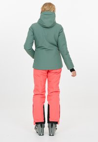 Skidjacka i grönt med huva, matchad med ljus korallfärgade byxor. Har svarta sidopaneler och grå skidstövlar. Slätt, vattentätt material.