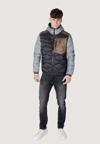 Polstret jakke med grå overdel og sort underdel, med en brun brystlomme, lynlåslukning og hætte. Sammen med mørke jeans og hvide sneakers.