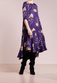 Fioletowa sukienka w kwiatowy wzór z rękawami 3/4 i rozkloszowanym dołem, w połączeniu z czarnymi, wysokimi kozakami za kolano. Gładki materiał o swobodnym kroju.