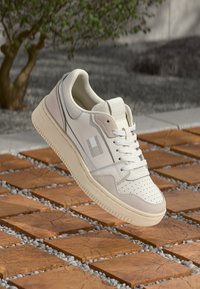 Weiße und beige Sneaker mit perforierter Zehenpartie und Schnürsenkeln, die über quadratische Holzdielen im Freien nahe einem Baum und Kies schwebt.
