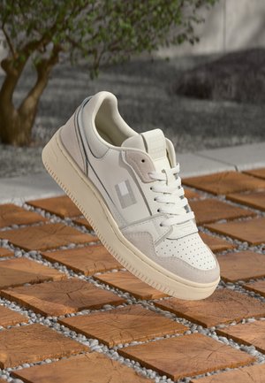 Sneaker bianco e beige con punta perforata e lacci, sospesa sopra piastrelle quadrate in legno all'aperto vicino a un albero e ghiaia.